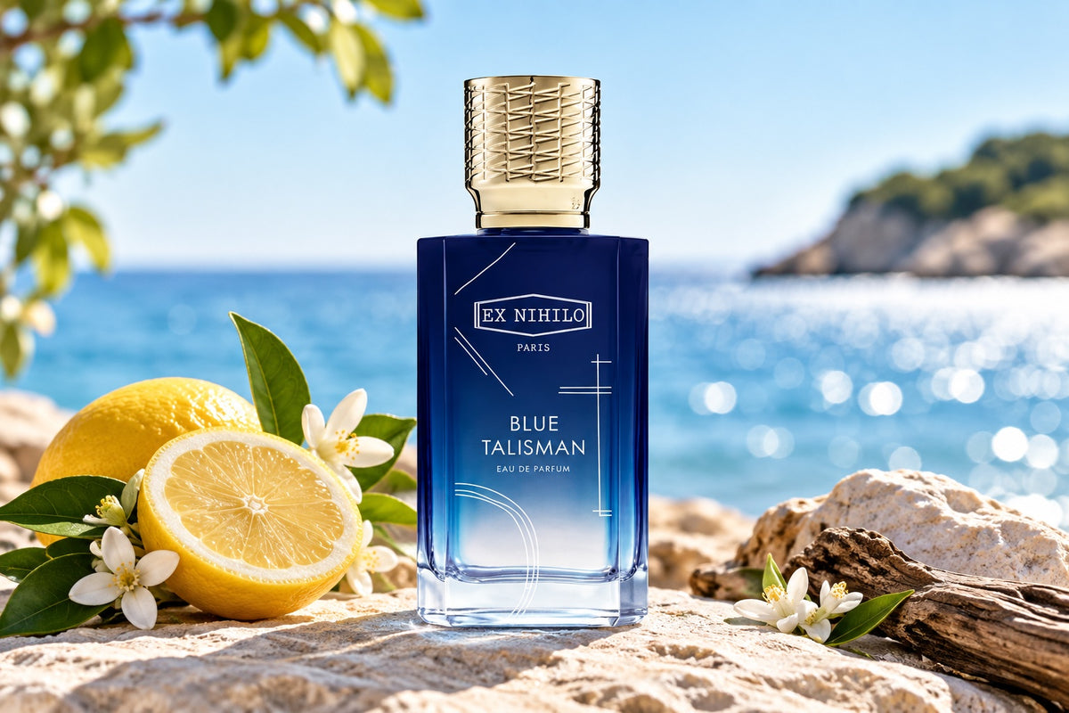 A bottle of Ex Nihilo Paris Blue Talisman Eau de Parfum stands on a beach setting with lemons, white flowers, rocks, and the blue ocean in the background. The bottle is dark blue with a textured gold cap.